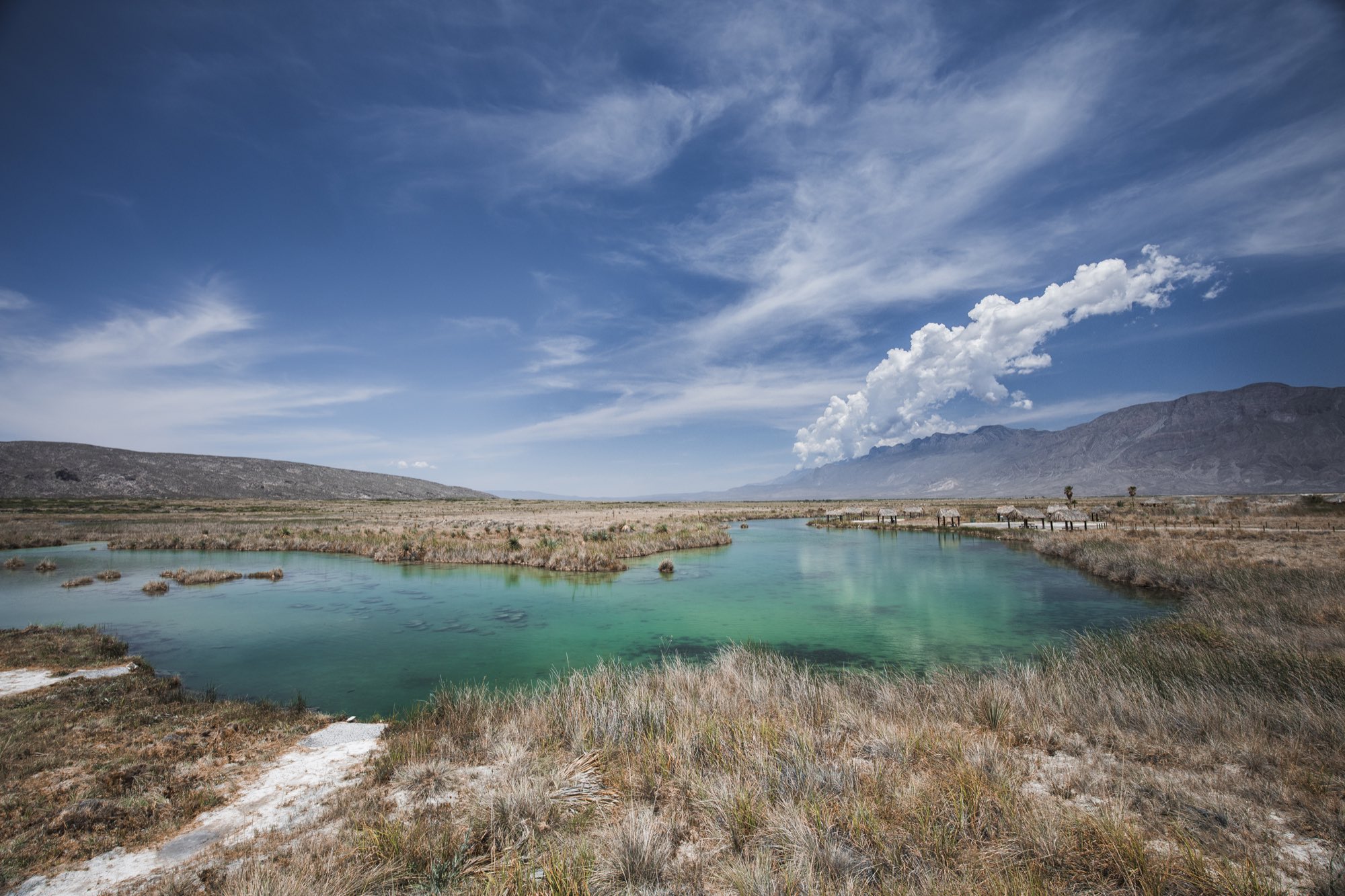 Humedal de agua turquesa con pastizal seco y sierra bajo cielo con nubes